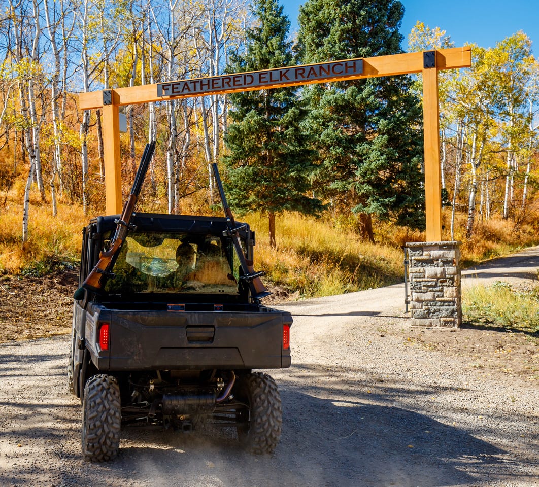 A UTV with rifles mounted on the back drives on a dirt road towards the entrance of Feathered Elk Ranch, surrounded by autumn trees.