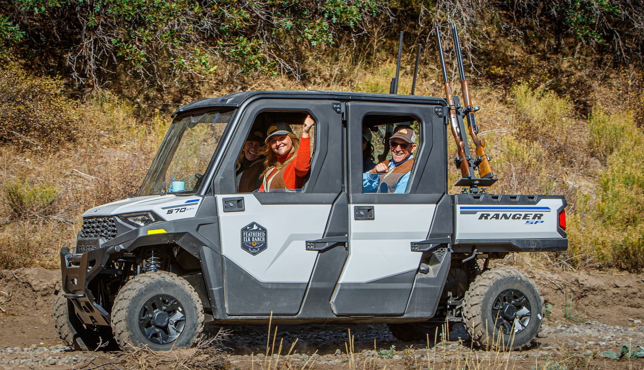 Two people in a UTV with rifles mounted on the back are driving through a brushy landscape.