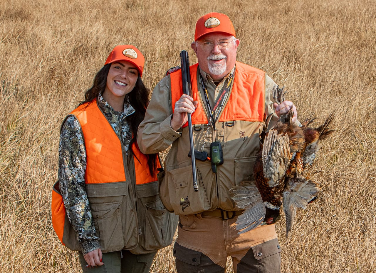 A man and a woman in hunting gear pose in a field, with the man holding a harvested ring-necked pheasant.