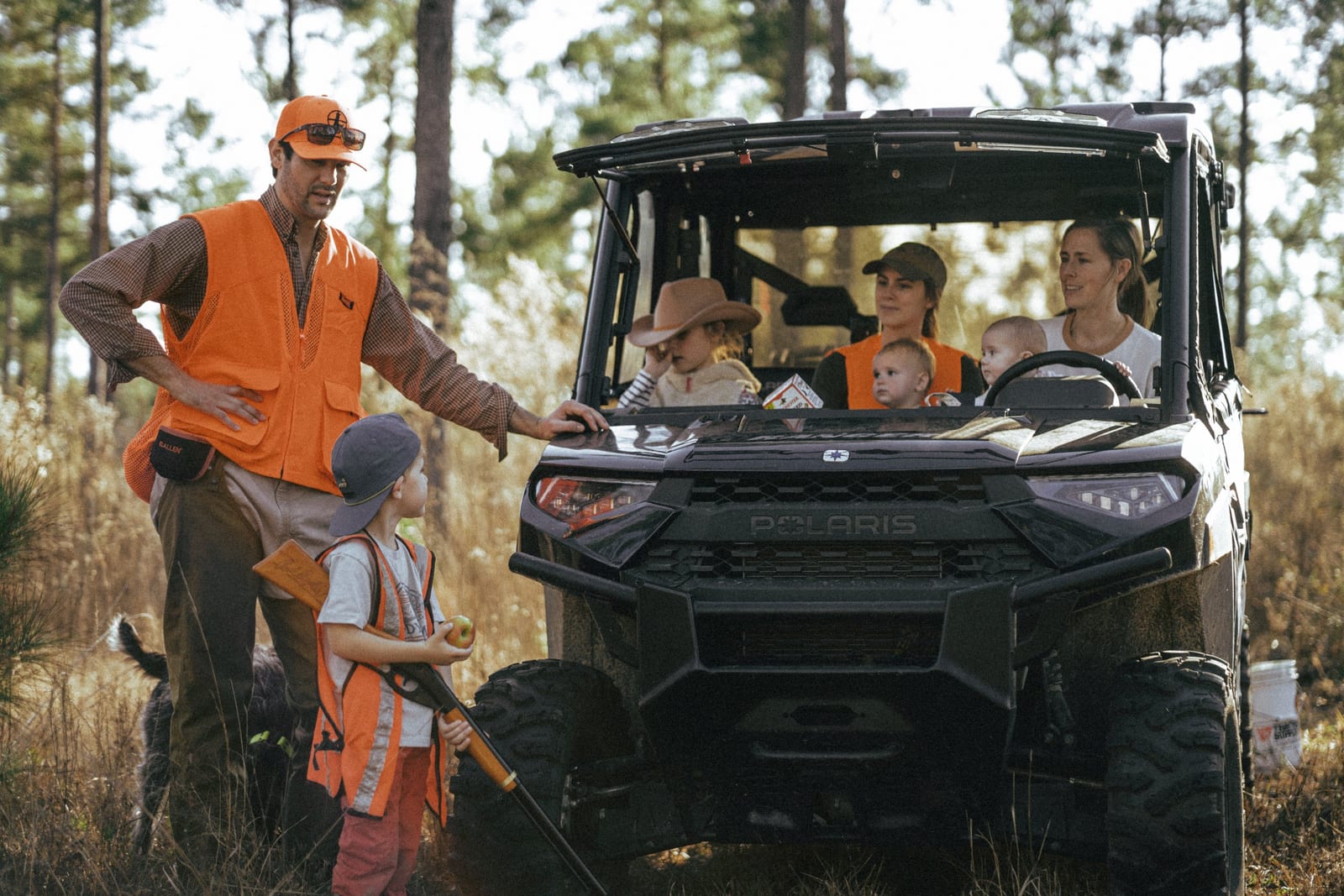 A man in an orange safety vest and a cap talks to children in safety vests near a black Polaris off-road vehicle in a wooded area.