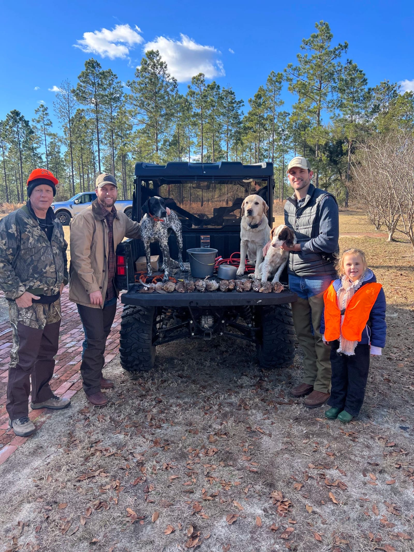 A group of five people and three dogs standing around a pickup truck with game birds laid out on the tailgate, in a wooded outdoor setting on a clear day.