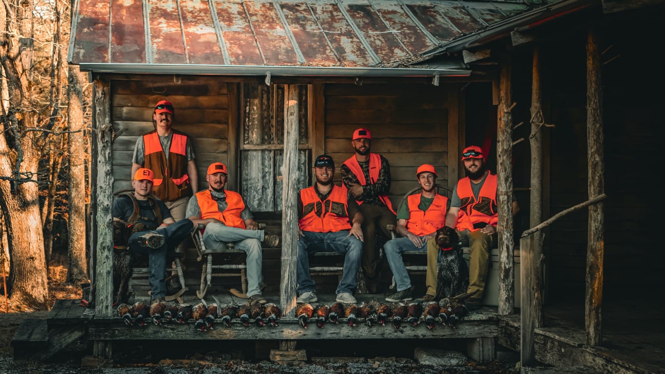 A group of hunters and a dog pose with their harvested ring-necked pheasants on a rustic porch.