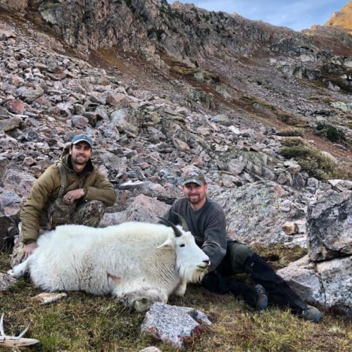Two hunters pose with a harvested mountain goat in a rocky, mountainous landscape.