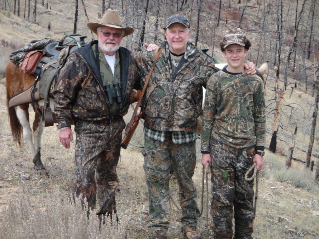 Three men in camouflage stand with a pack mule in a wooded, hilly area.