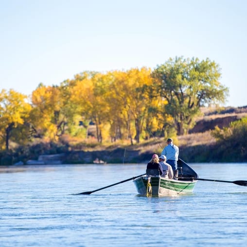Three people are in a green drift boat on a river, with colorful autumn trees lining the far bank.