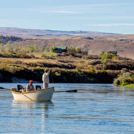 Two men in a drift boat on a river, with one man fly fishing, against a backdrop of rolling hills and distant buildings.