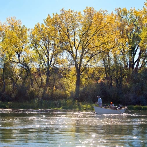 Two people in a boat are fishing on a river lined with trees displaying autumn foliage.