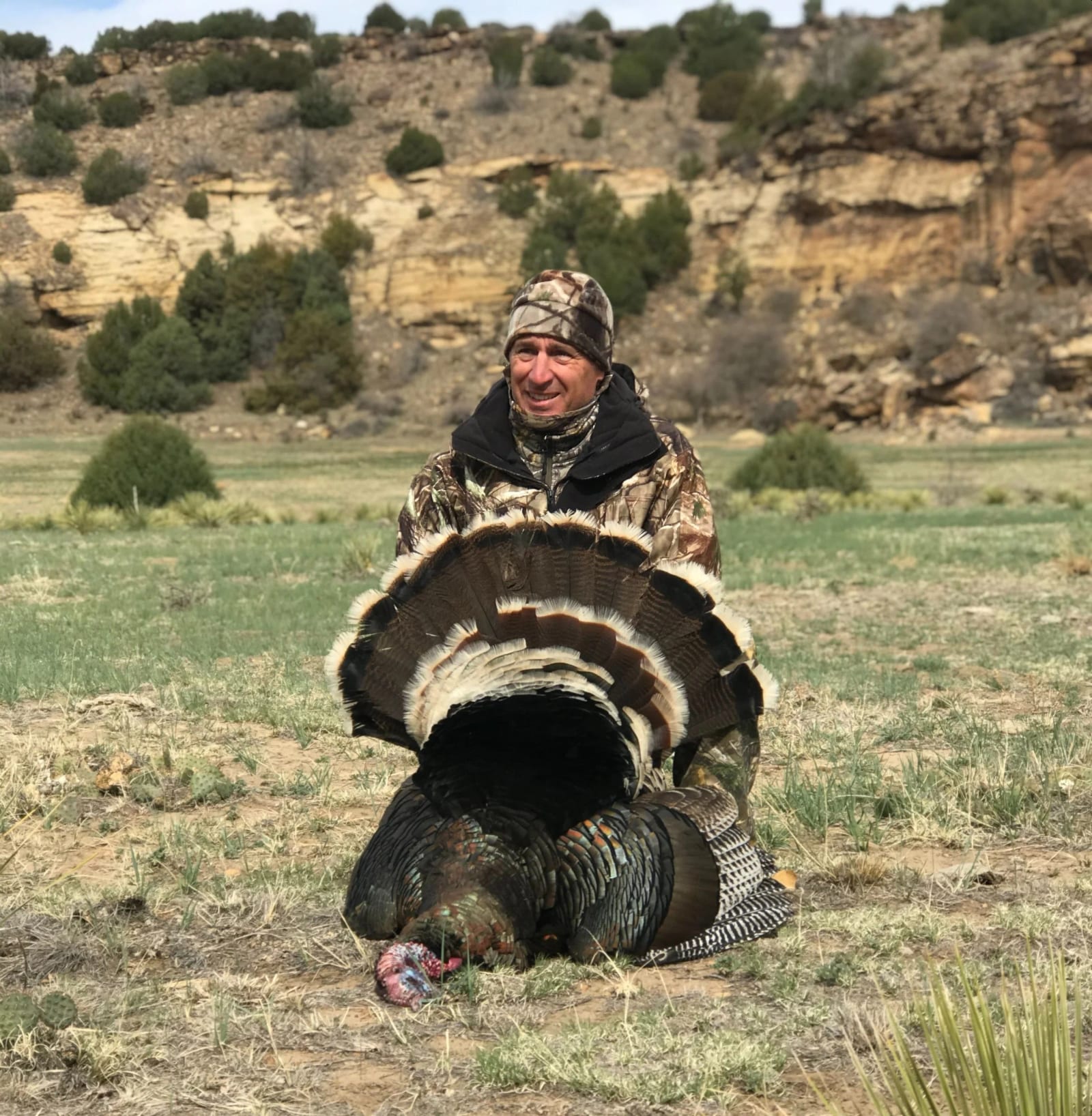 A hunter poses with a harvested wild turkey in a field.