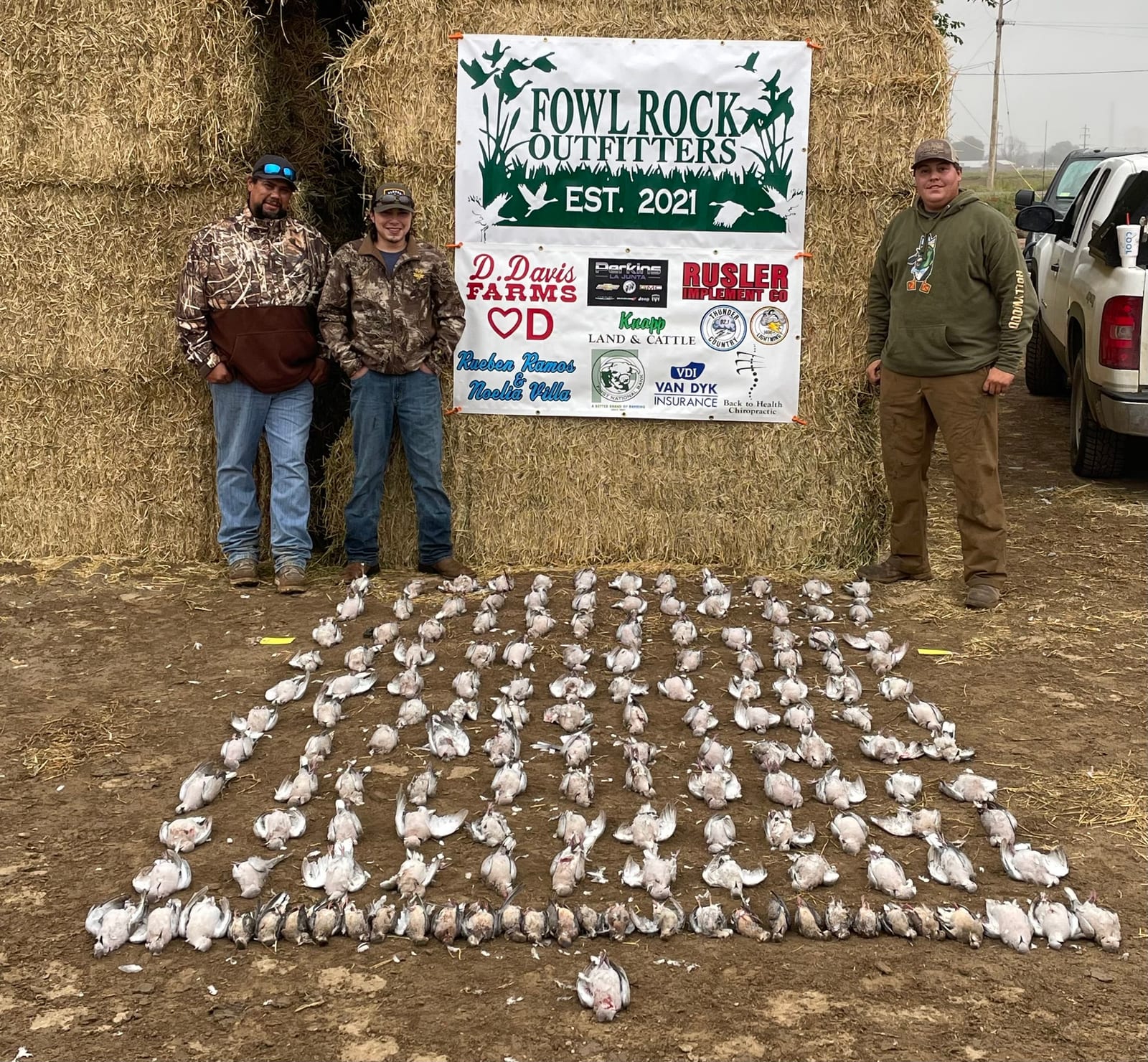 Three hunters pose with a large harvest of mourning doves laid out on the ground in front of hay bales and a Fowl Rock Outfitters banner.