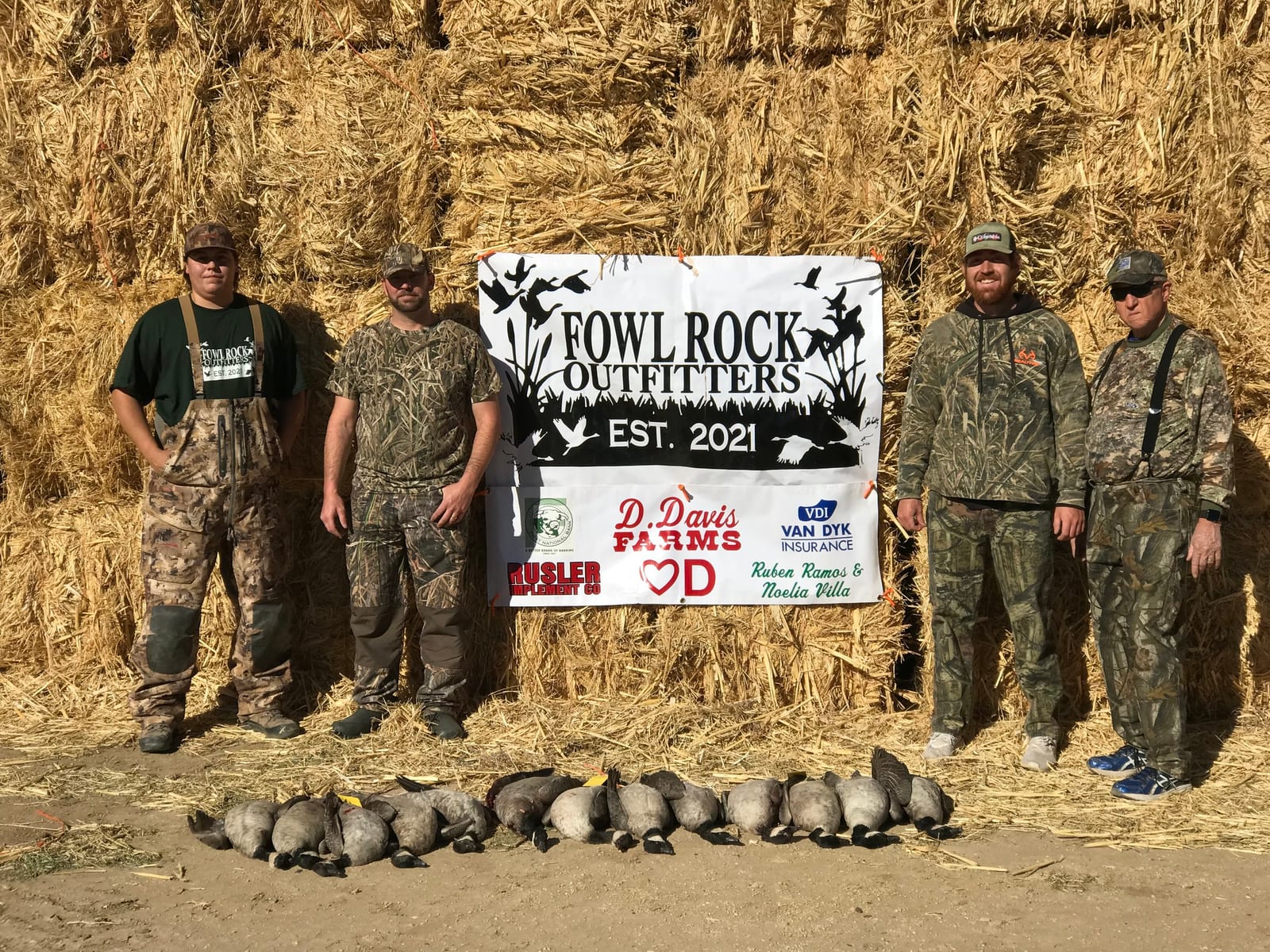 Four hunters pose with their harvested Canada geese in front of a hay bale wall and an outfitter banner.