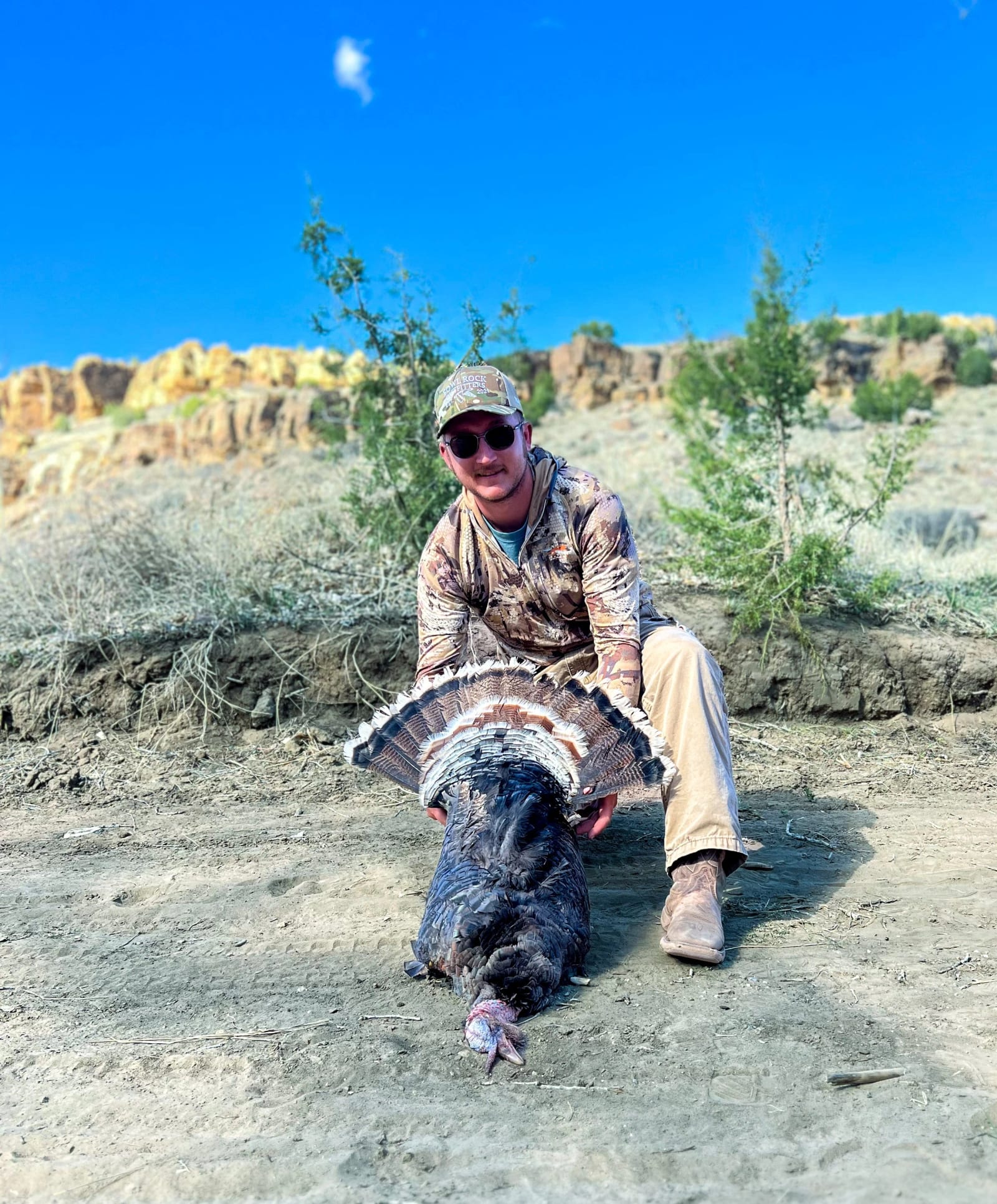 A hunter poses with a harvested wild turkey in a field.
