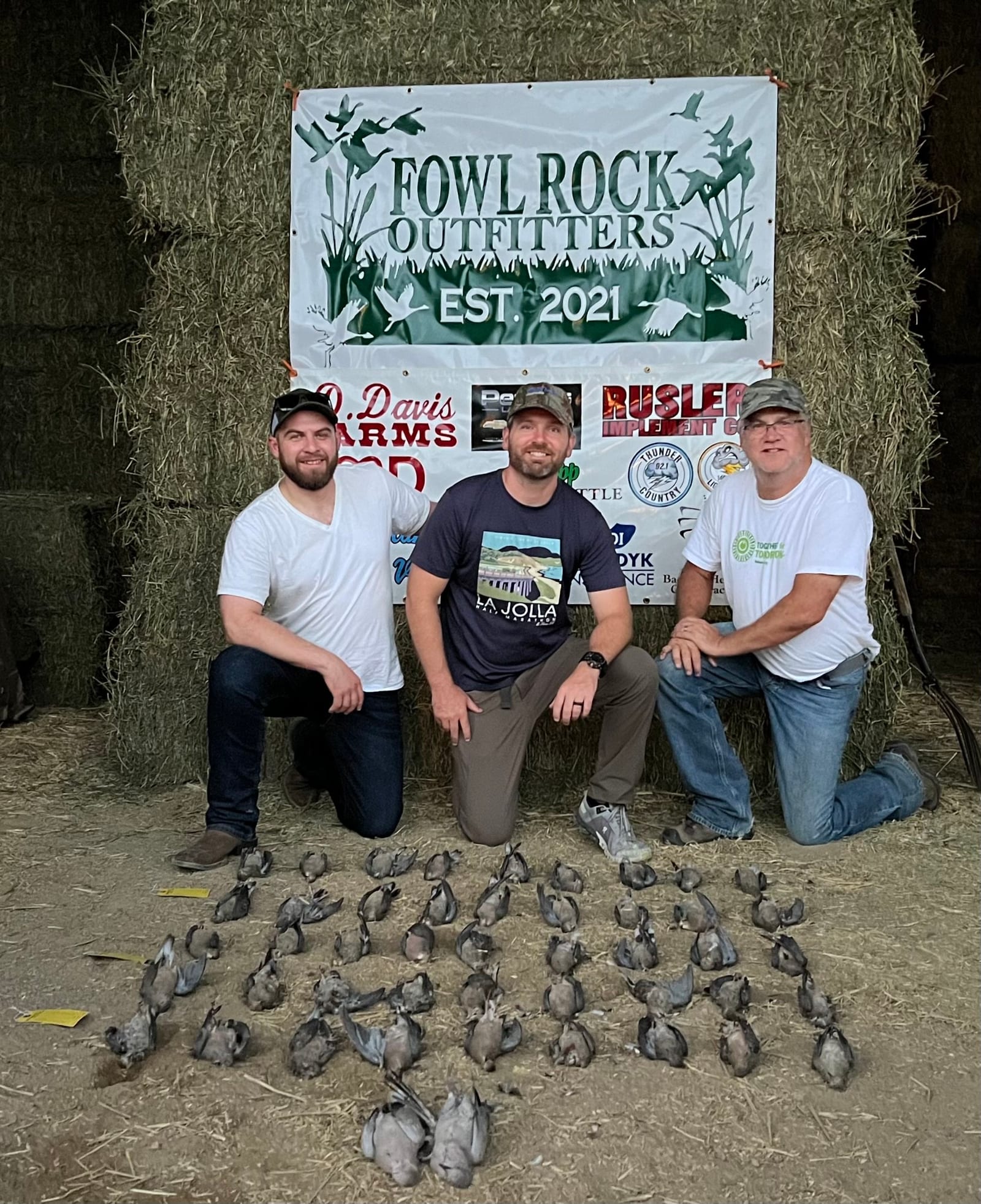 Three men pose with a large harvest of mourning doves in front of a Fowl Rock Outfitters banner.