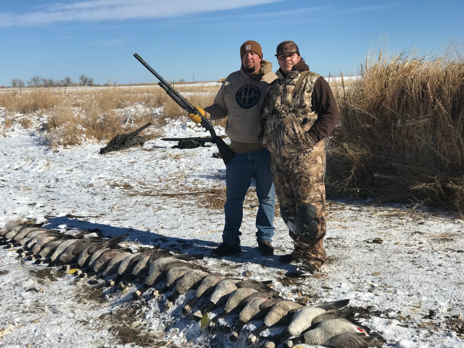 Two hunters pose with a line of harvested Canada geese in a snowy field.
