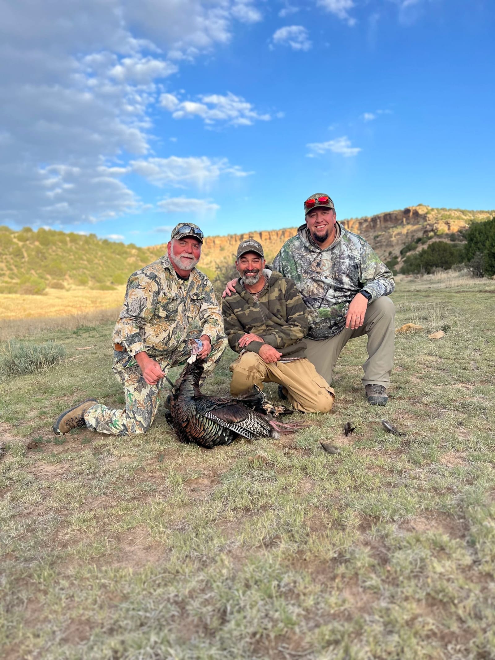 Three hunters pose with a harvested wild turkey in a field under a blue sky.