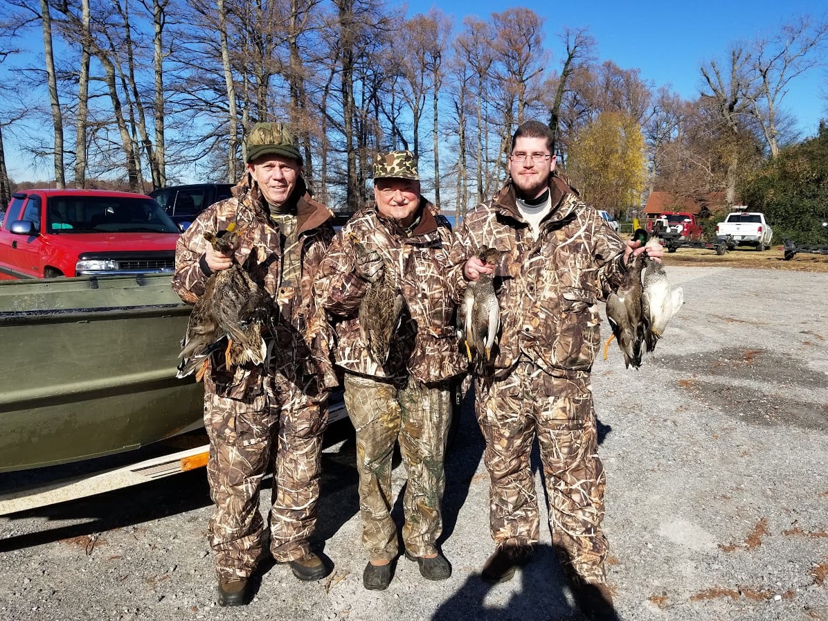 Three hunters in camouflage stand next to a boat, holding harvested ducks.