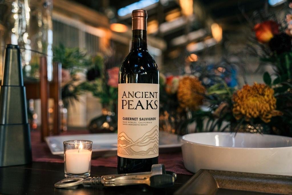 A bottle of Ancient Peaks Cabernet Sauvignon wine, a lit candle, and a corkscrew are on a table.
