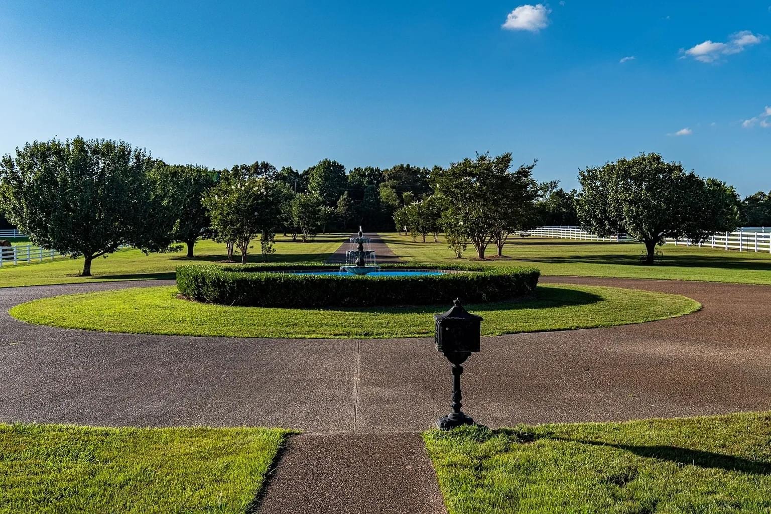 A circular driveway with a fountain in the center, surrounded by green grass and trees under a clear blue sky.