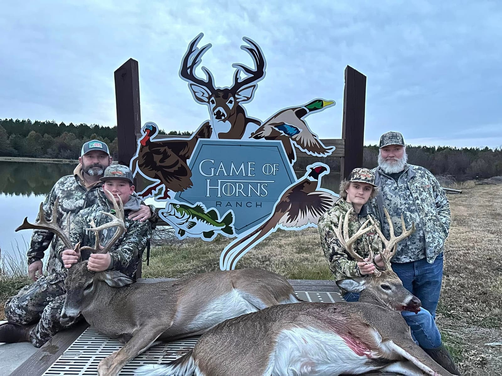 Two men and a boy pose with two harvested whitetail deer in front of a Game of Horns Ranch sign.