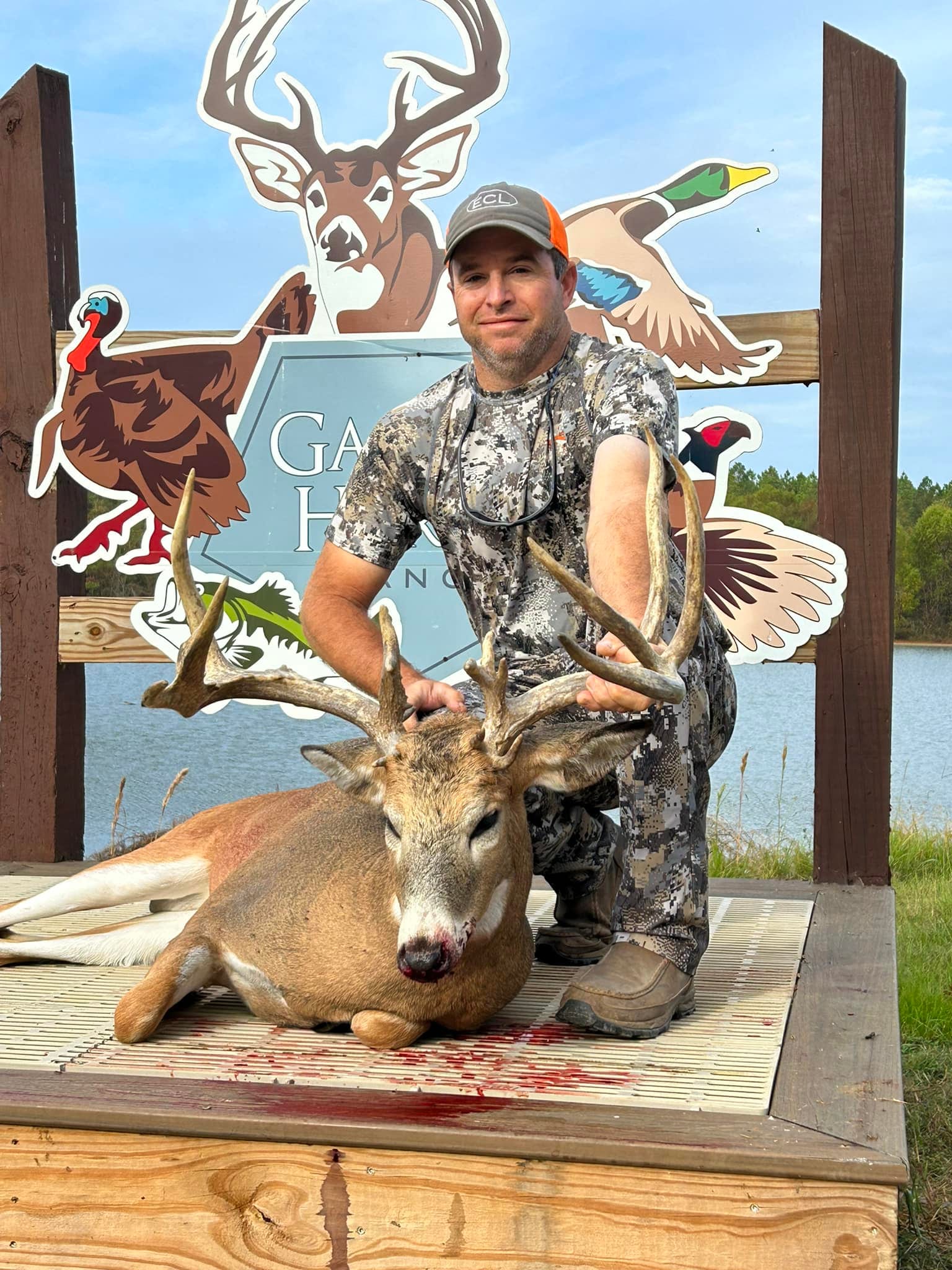 A hunter kneels next to a harvested whitetail deer on a wooden platform, with a lake and graphic cutouts in the background.