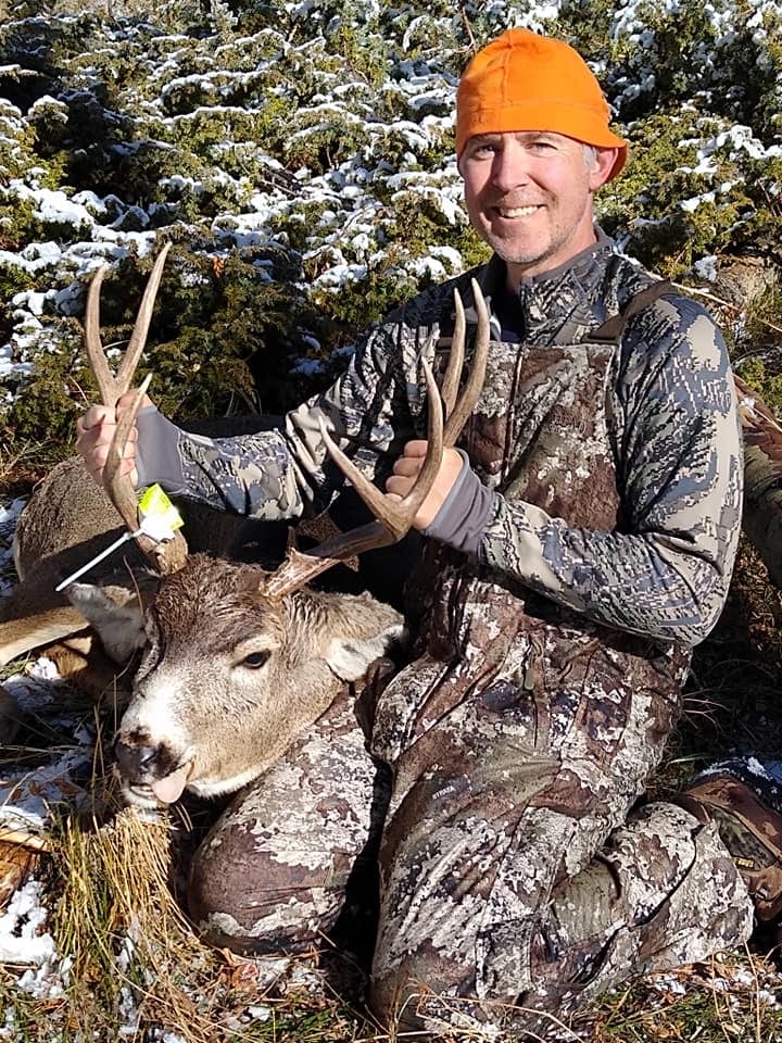 A hunter in camouflage and an orange hat poses with a harvested mule deer in a snowy, wooded area.