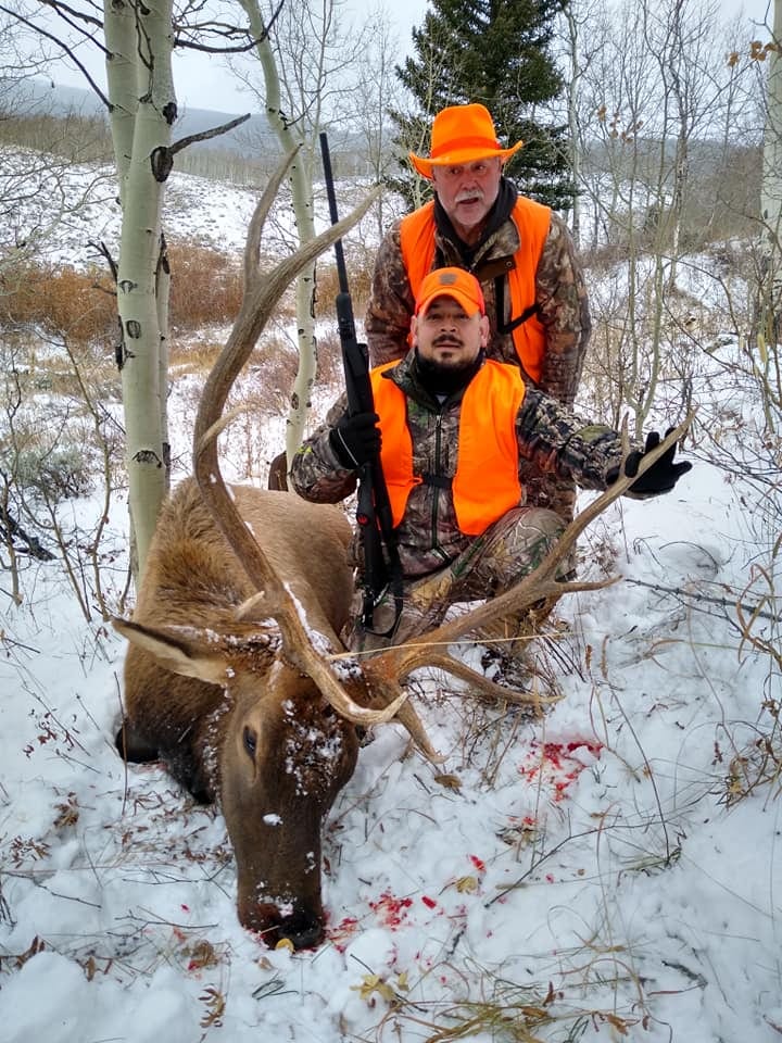 Two hunters pose with a harvested elk in a snowy, wooded area.