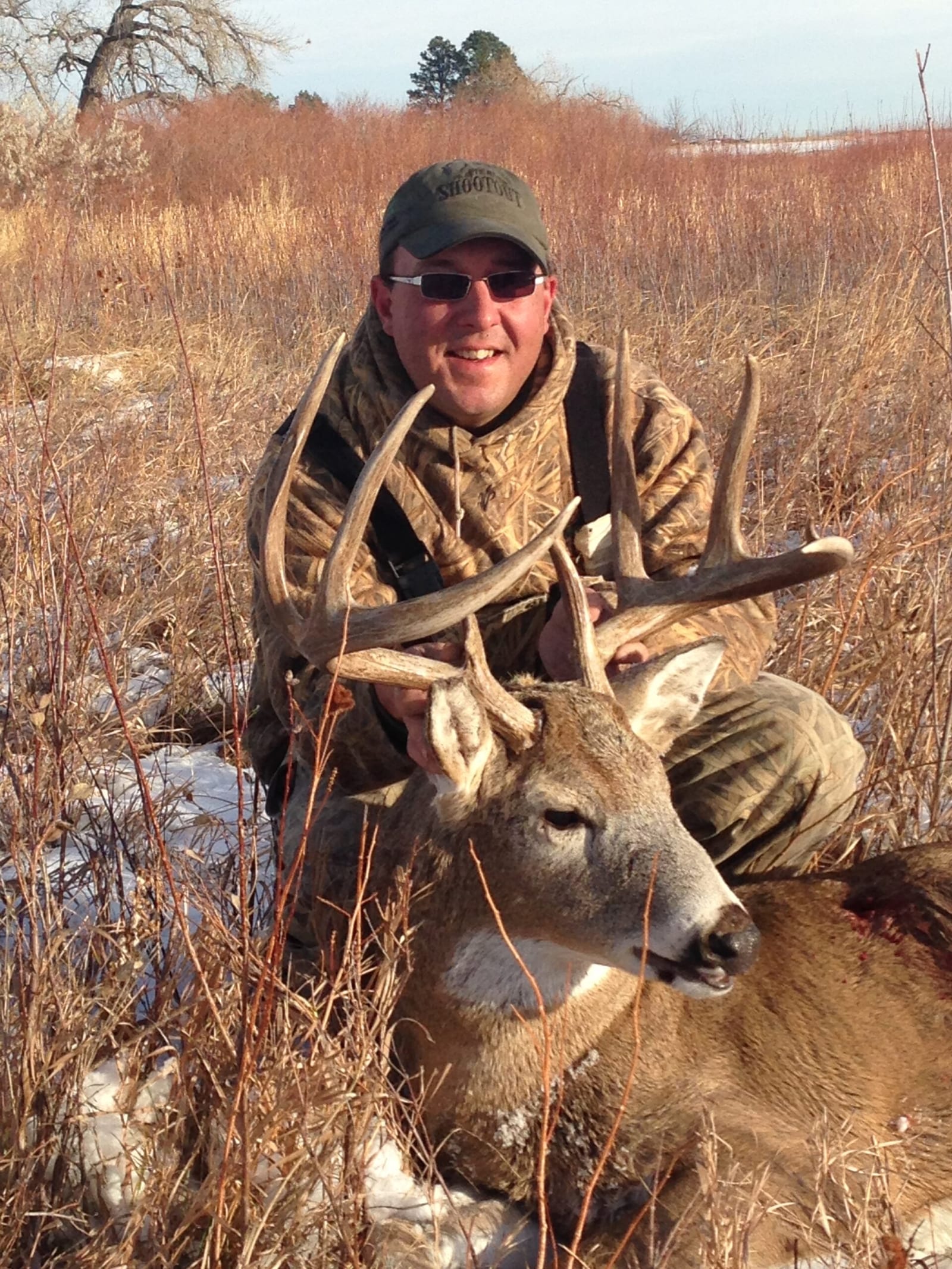 A hunter in camouflage poses with a harvested whitetail deer in a field with dry grass and some snow.