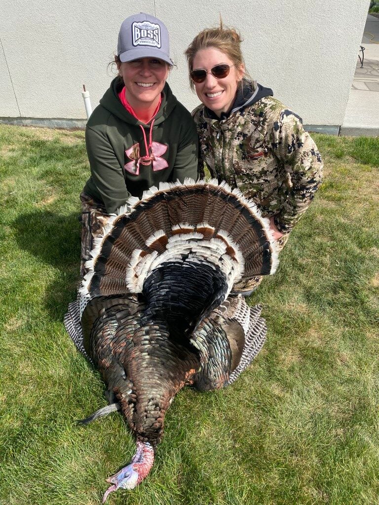 Two women pose with a harvested wild turkey on a grassy lawn.