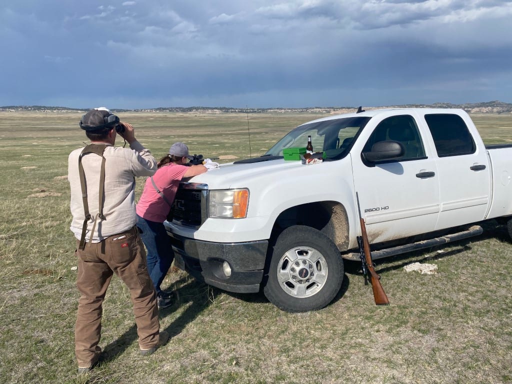 A man looks through binoculars while a woman leans on a white pickup truck with a rifle leaning against it in a grassy field under a cloudy sky.