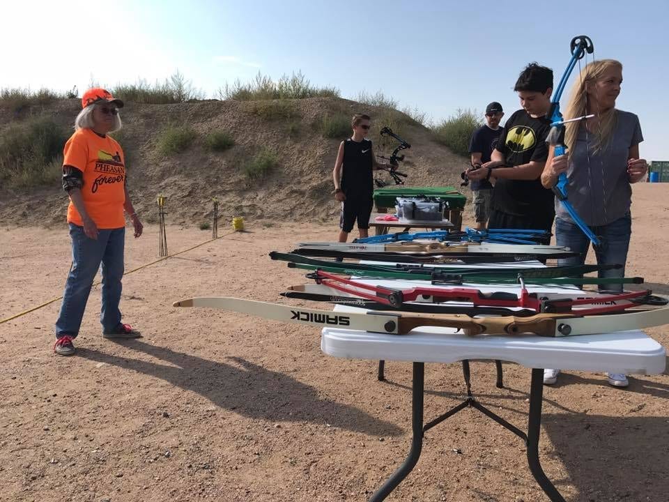 People are at an outdoor archery range with various bows on a table.