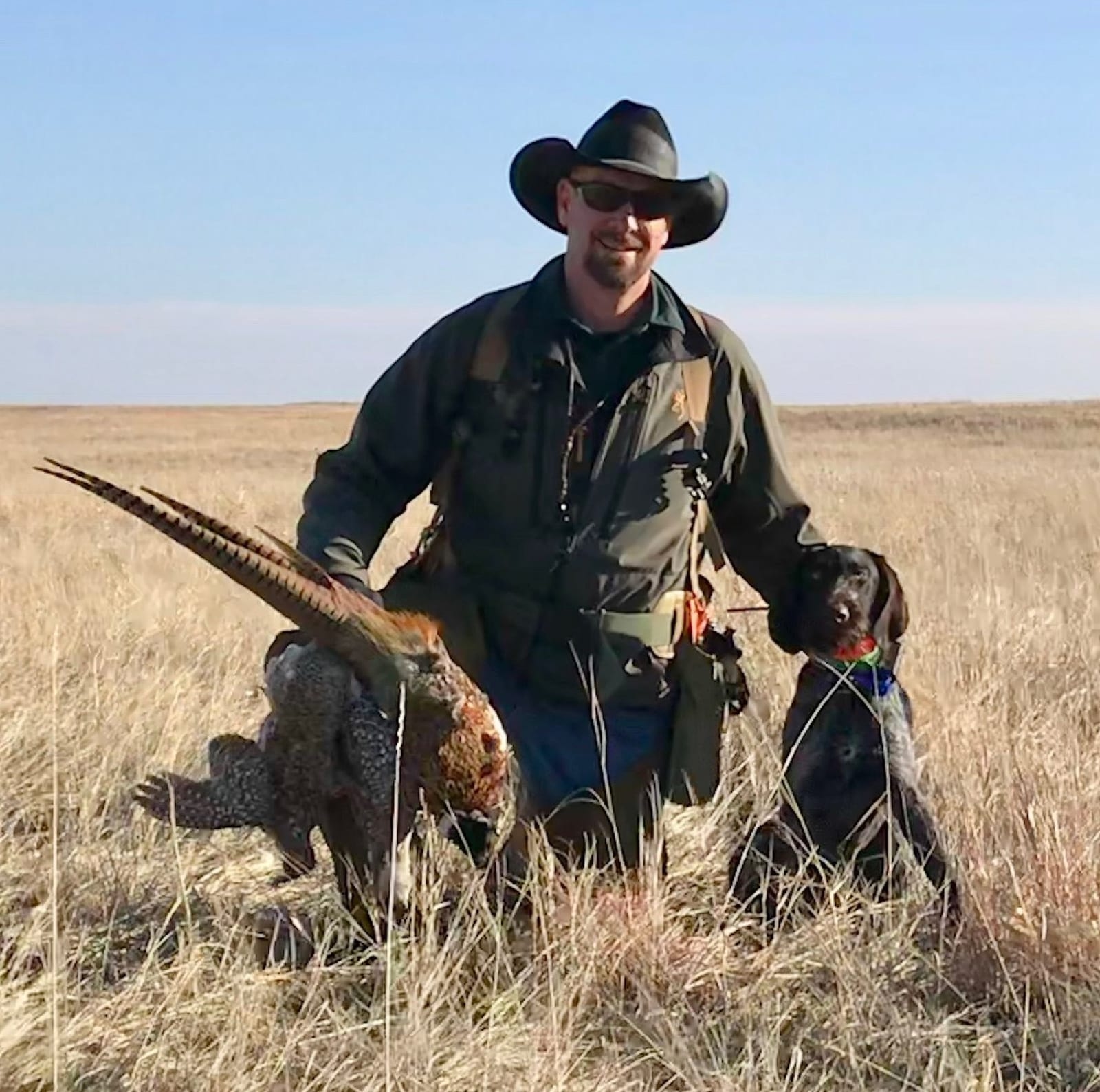 A hunter poses with a harvested ring-necked pheasant and a hunting dog in a field.