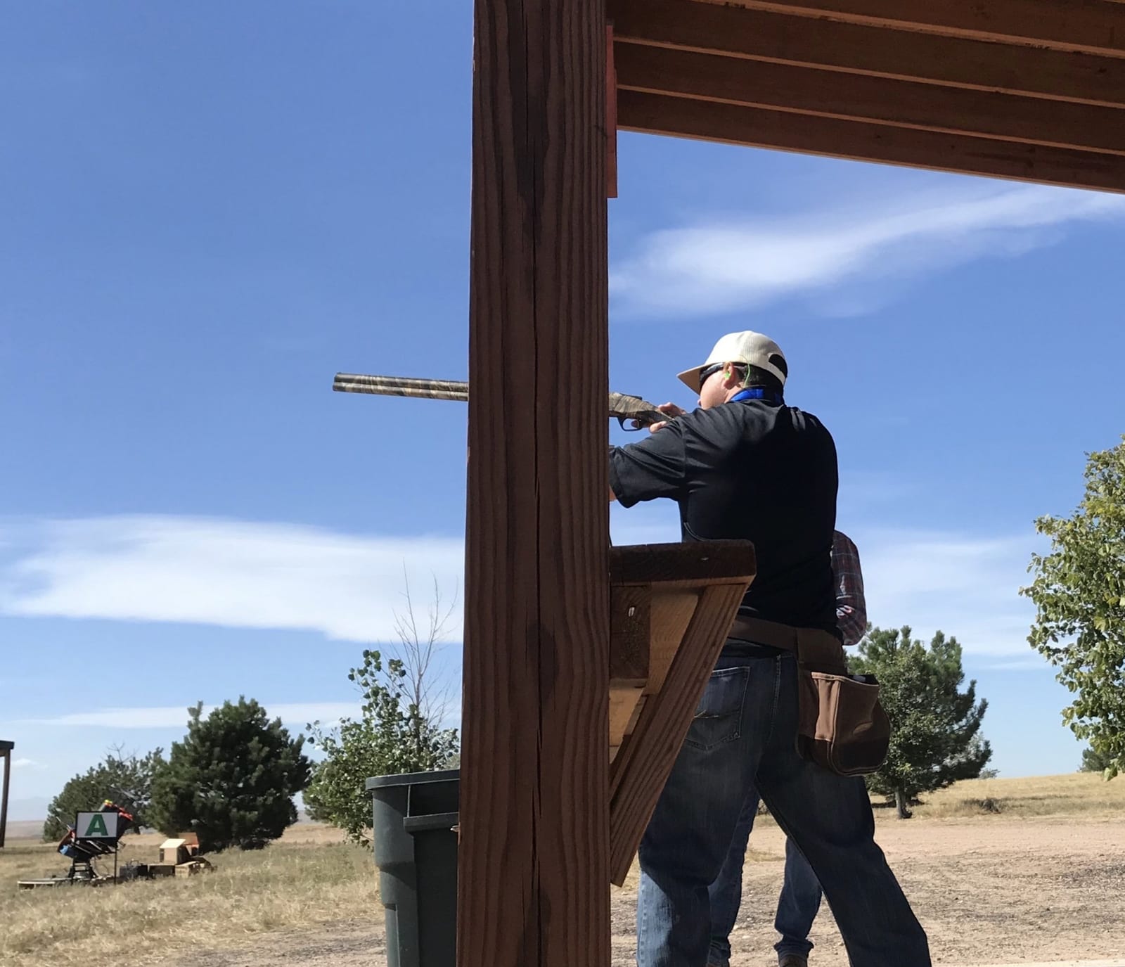 A person in a hat and dark shirt aims a shotgun at a shooting range under a blue sky.