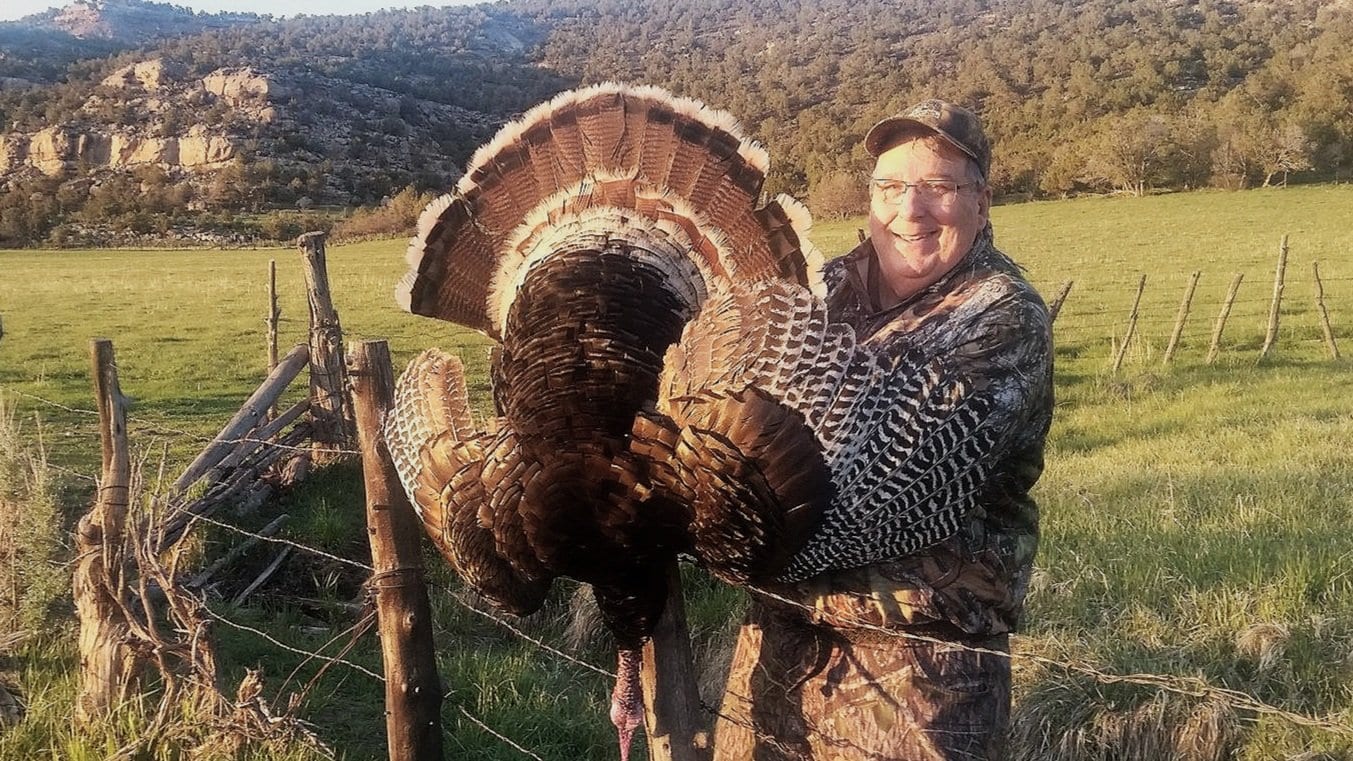 Hunter holding a turkey in a rural landscape with hills and trees.