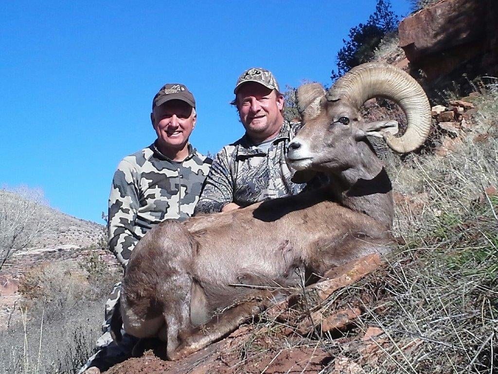 Two men in camouflage clothing posing with a bighorn sheep on rocky terrain, under a clear blue sky.