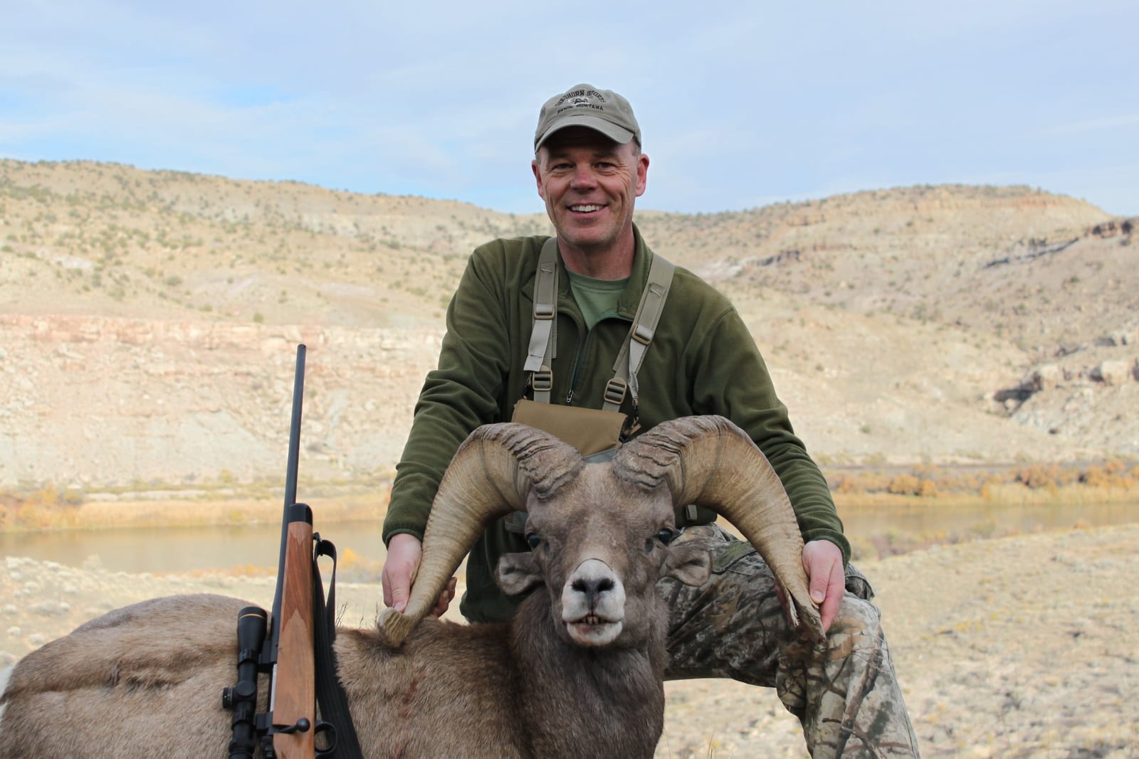 Man in outdoor gear posing with a bighorn sheep against a mountainous landscape.