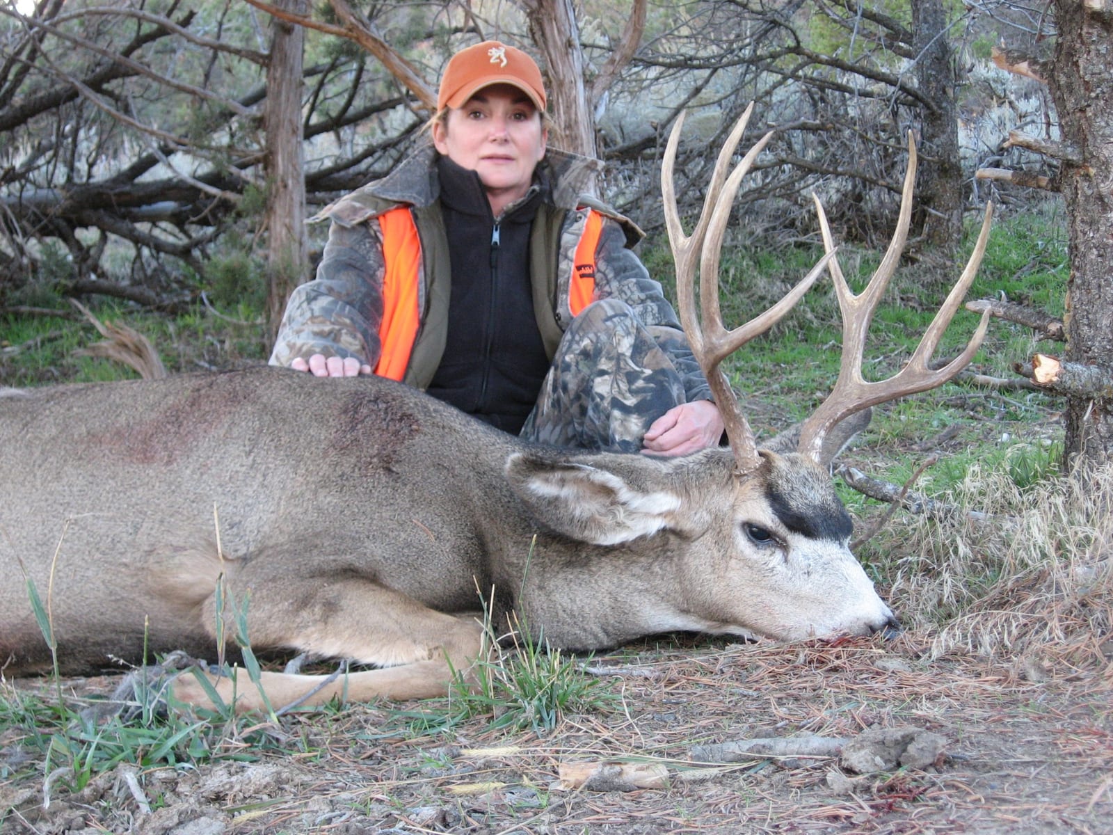 Hunter posing with harvested deer, wearing camouflage outfit and orange cap, in wooded area.