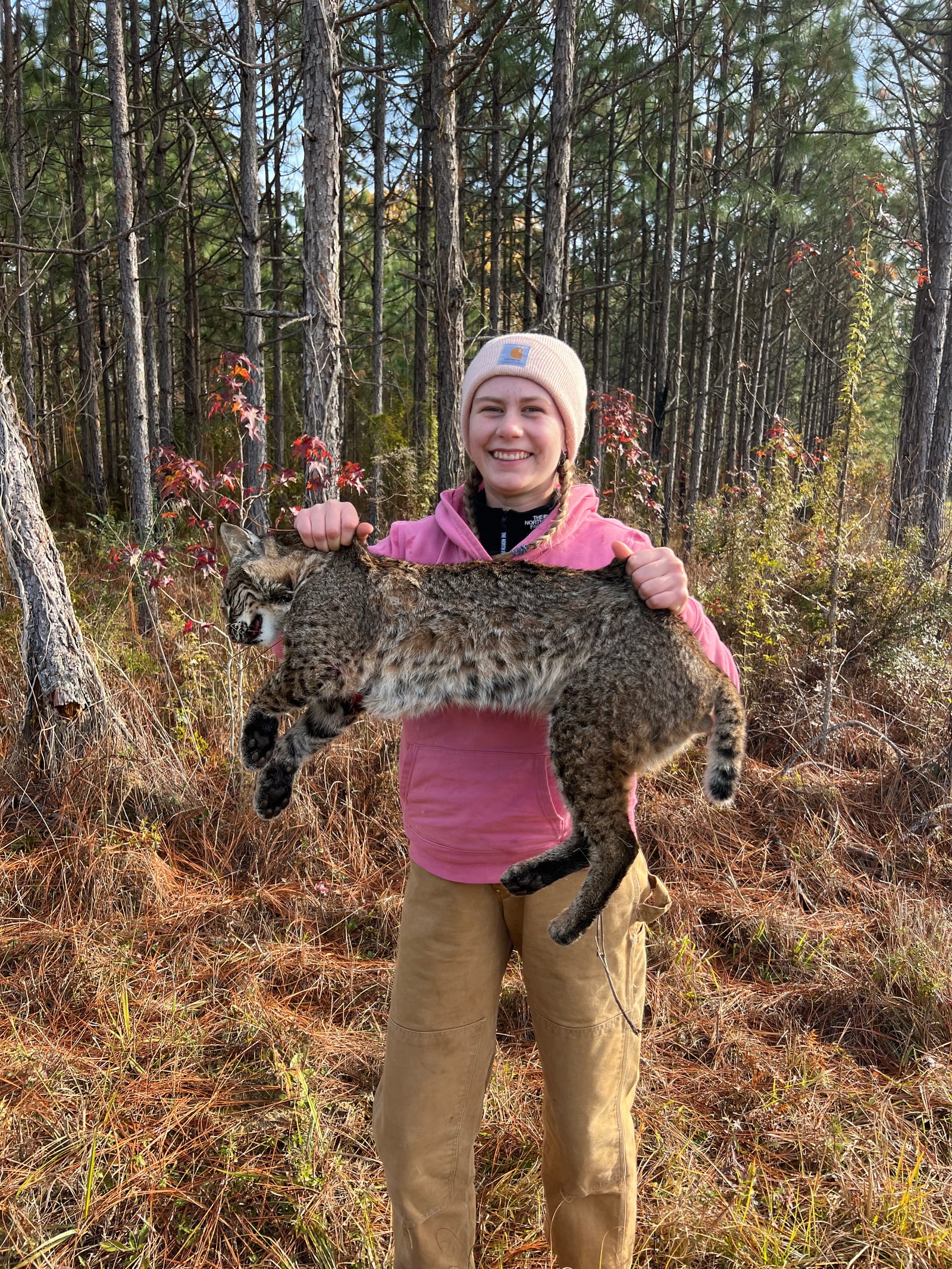 A young woman in a pink hoodie and a beanie holds a harvested bobcat in a forest.