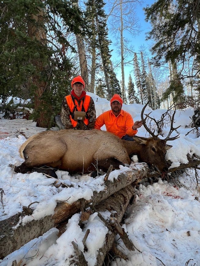 Two hunters in orange safety gear sitting behind a dead elk with large antlers in a snowy forest setting. 3rd rifle elk hunt in GMU 044 Gypsum Colorado. Guided elk hunts Colorado.