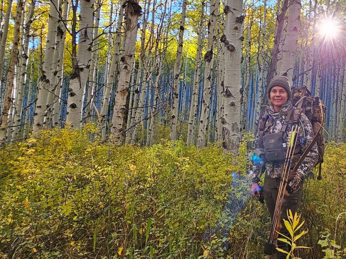 A woman hiking in a forest of aspen trees with sunlight shining through the trees. Guided elk hunt with recurve bow in GMU 044, Colorado. Eagle County.