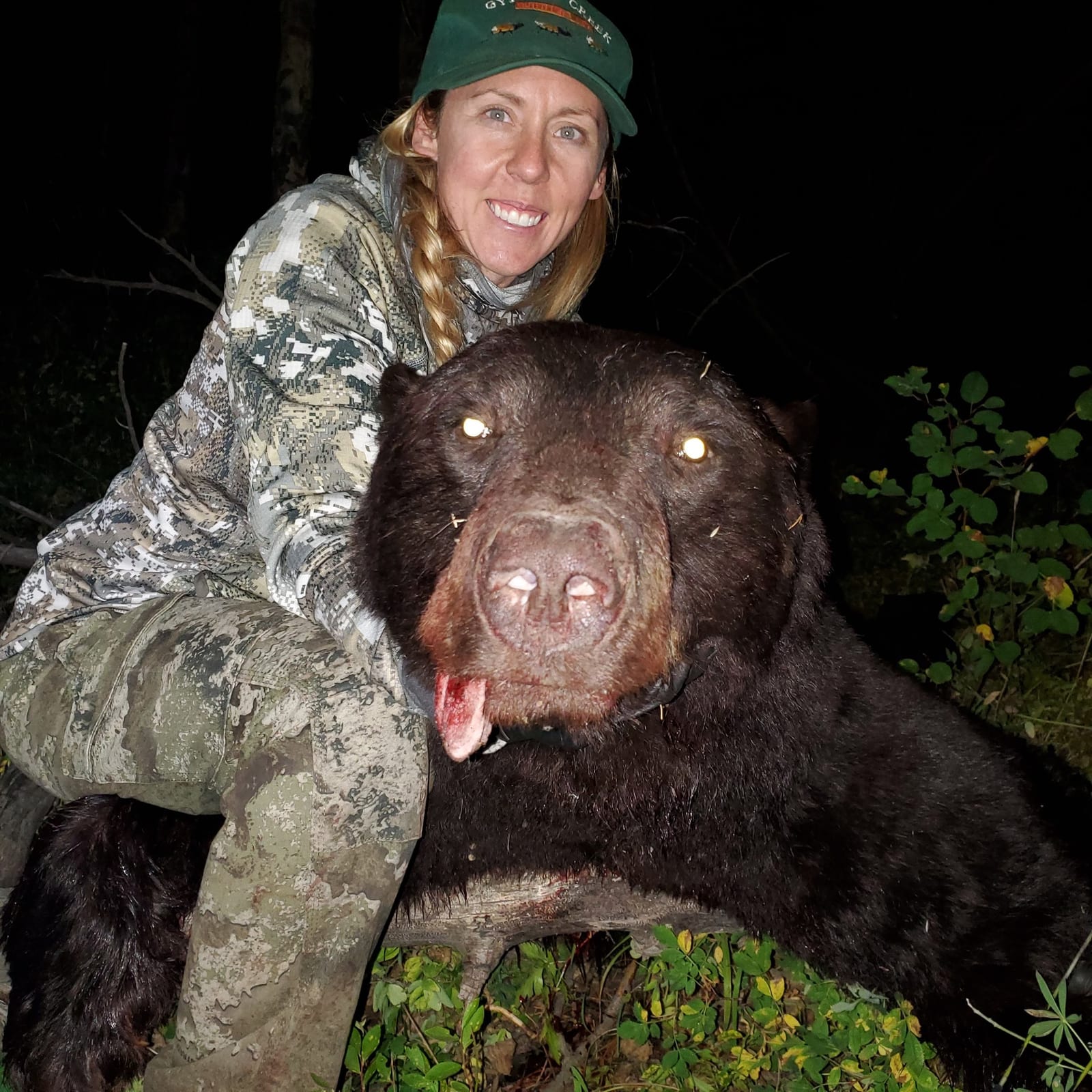 A woman dressed in camouflage clothing and a green hat posing with a large, dead black bear in a forest at night. Record black bear taken in GMU 044 in Colorado.