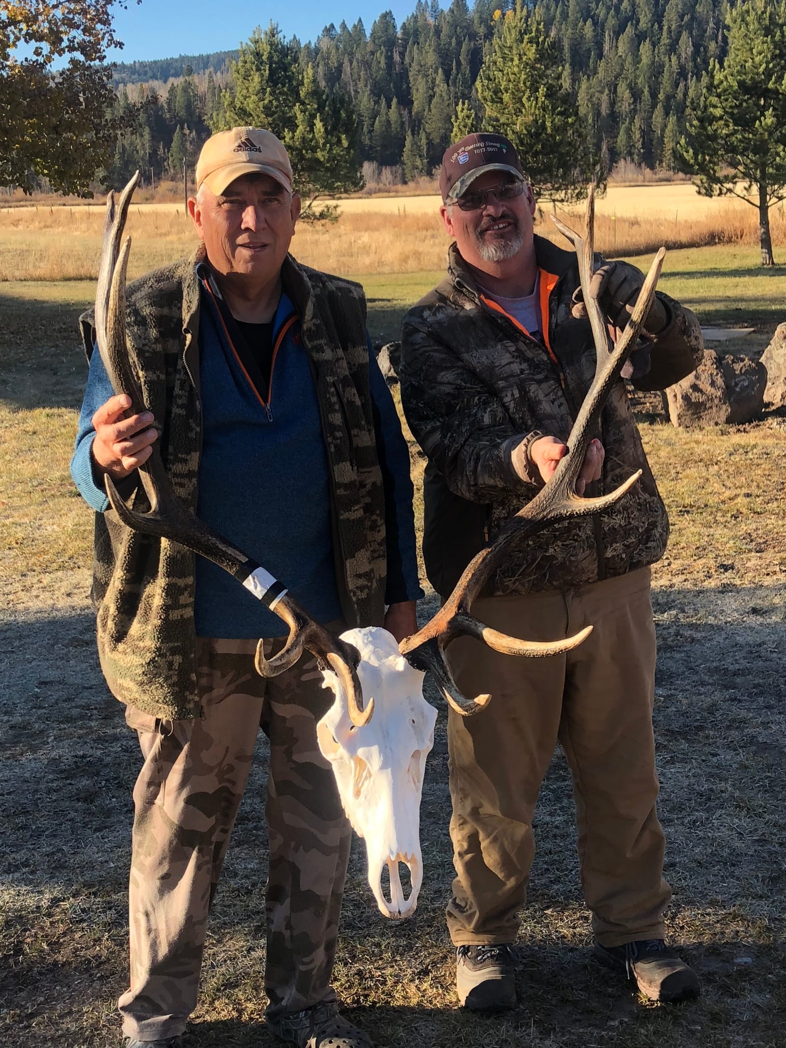 Two men pose with a mule deer skull and antlers in a field.