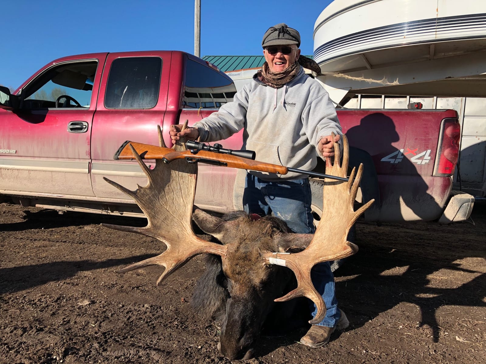 A hunter poses with a harvested moose and a rifle in front of a red truck.