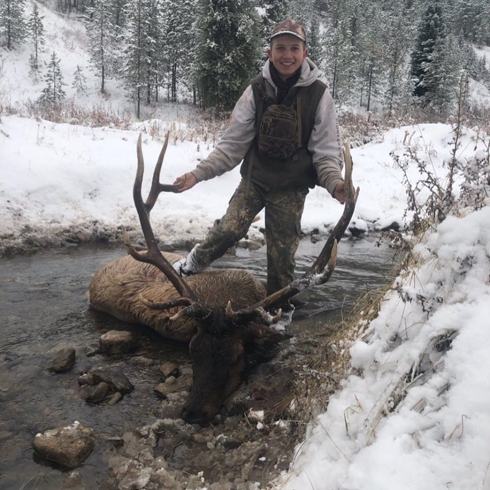 A hunter poses with a harvested elk in a snowy stream.
