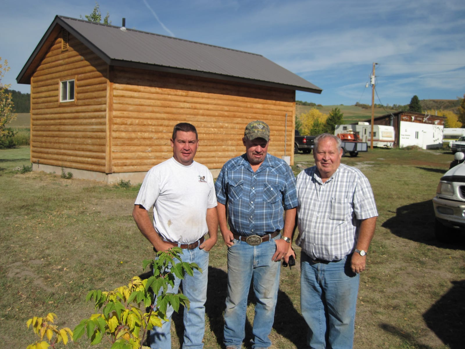 Three men stand in front of a log cabin-style building on a sunny day.