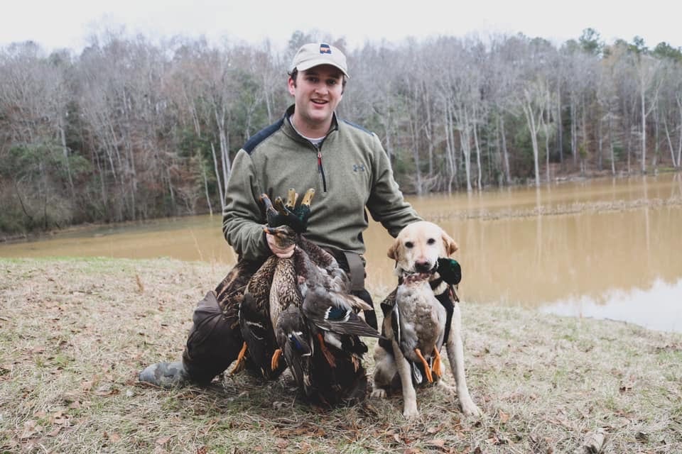 A man and a dog pose with harvested mallard ducks by a body of water.