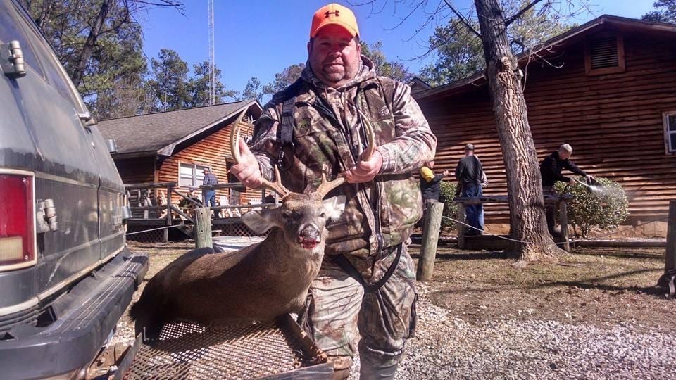 A hunter in camouflage poses with a harvested whitetail deer in front of a lodge.