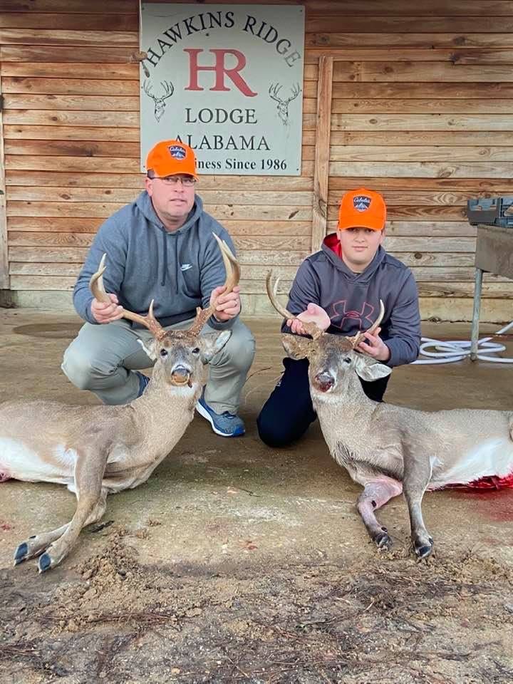 Two men pose with two harvested whitetail deer in front of a wooden lodge building.