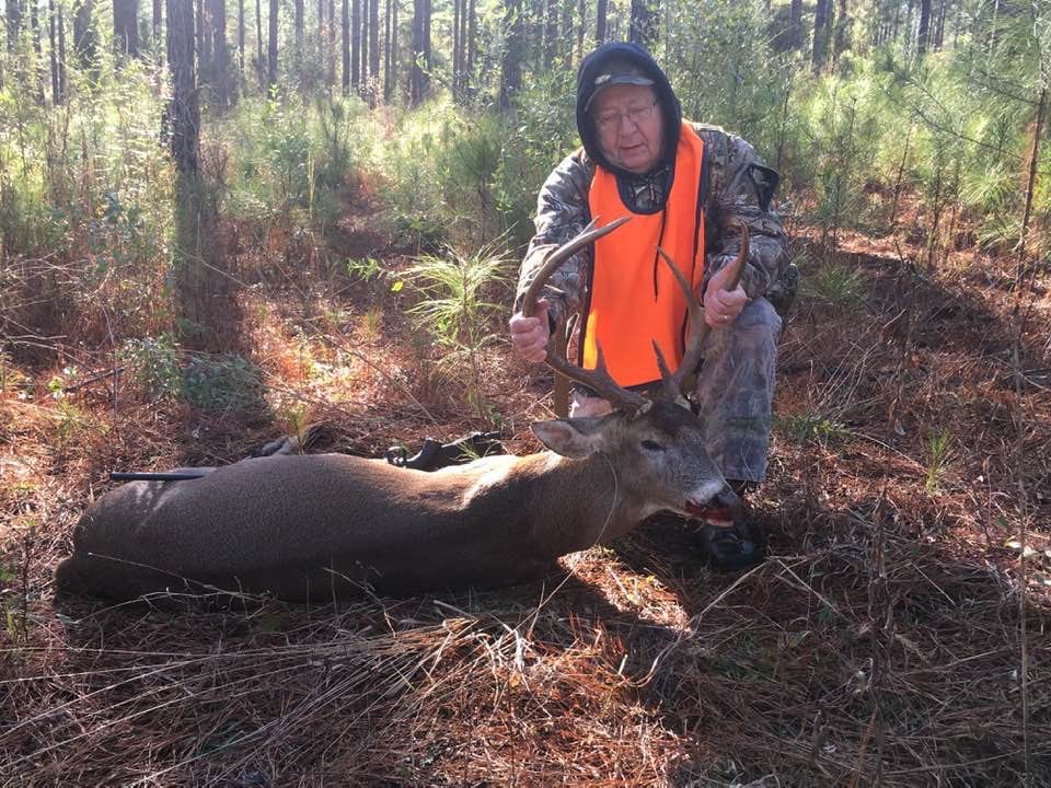 A hunter in camouflage and an orange vest kneels next to a harvested whitetail deer in a forest.
