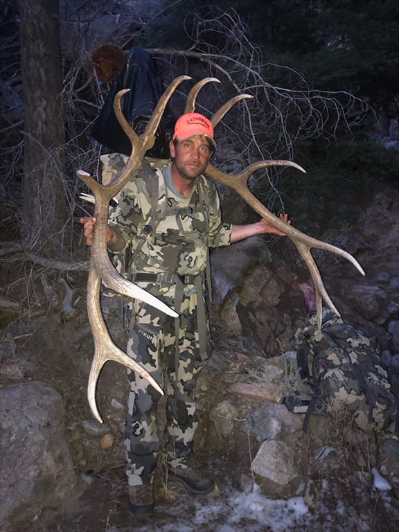 A hunter in camouflage poses with a harvested elk in a rocky, wooded area.