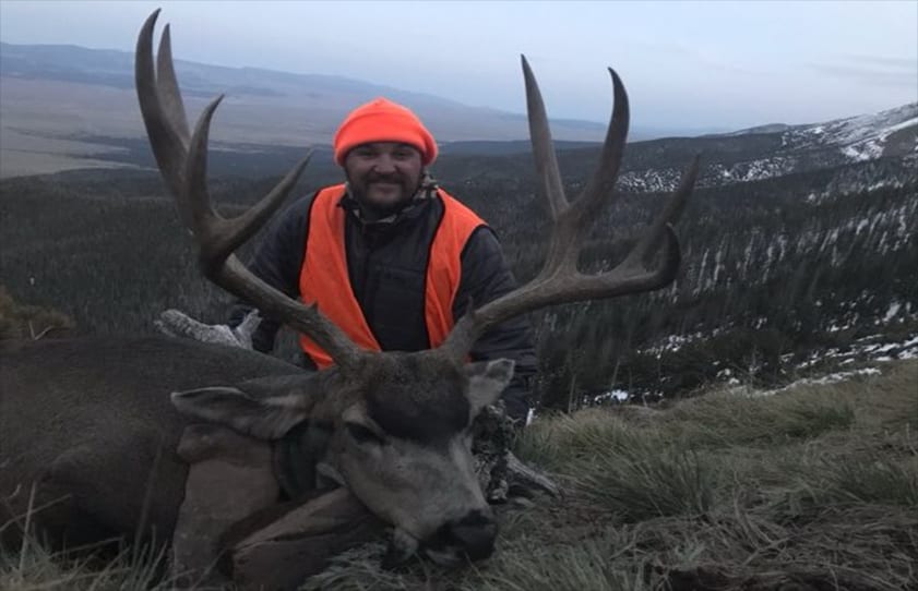 A hunter in an orange hat and vest poses with a harvested mule deer in a mountainous, snowy landscape.
