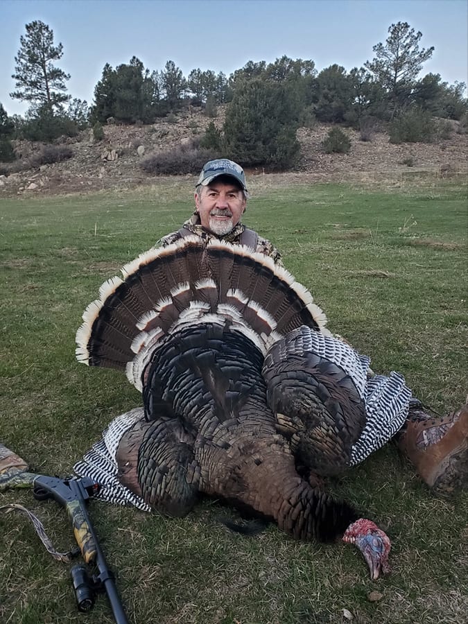 A hunter poses with a harvested wild turkey in a grassy field.